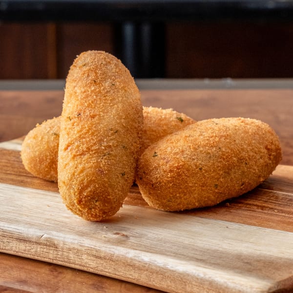 Three golden-brown, breaded croquettes, likely from John's Deli, rest on a wooden cutting board.
