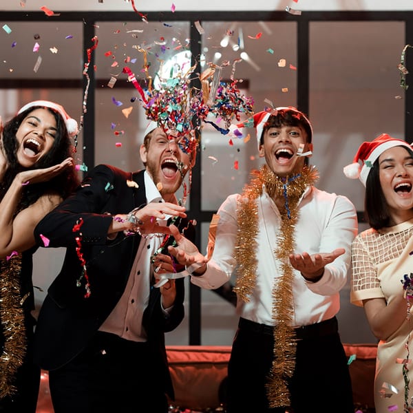 Four joyful people in Santa hats and tinsel celebrate with colorful confetti falling around them.