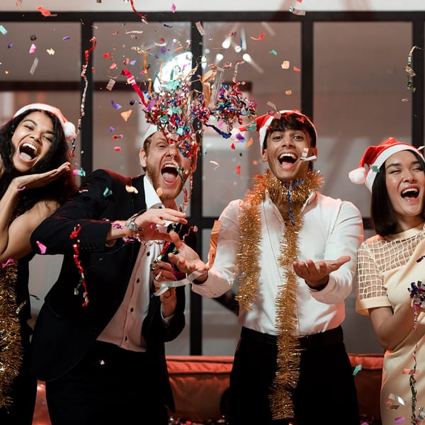 Four joyful people in Santa hats and tinsel celebrate with colorful confetti exploding around them.
