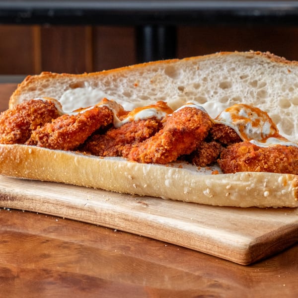 A John's Deli sandwich with crispy chicken, white sauce, and hot sauce on a wooden board.