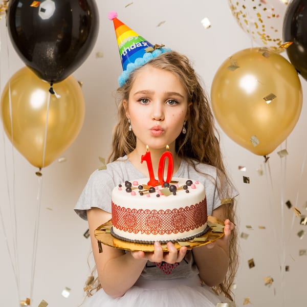 Young girl in a party hat holding a birthday cake with "10" candles, surrounded by balloons.