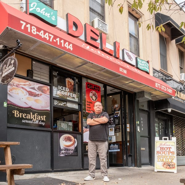 A man stands proudly in front of John's Deli, featuring a red awning and window advertisements.