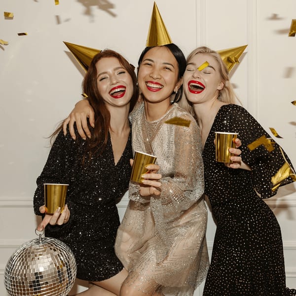 Three women in party hats and sequined dresses laughing, holding gold cups, with confetti falling.