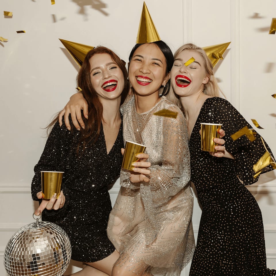 Three women in party hats and sequined dresses laughing, holding gold cups, with confetti falling.